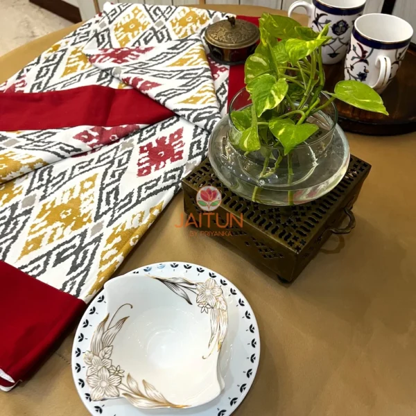 Ikat Harmony bed sheet featuring maroon edges and mustard ikat motifs, styled with green plant and classic tableware for a decor view.